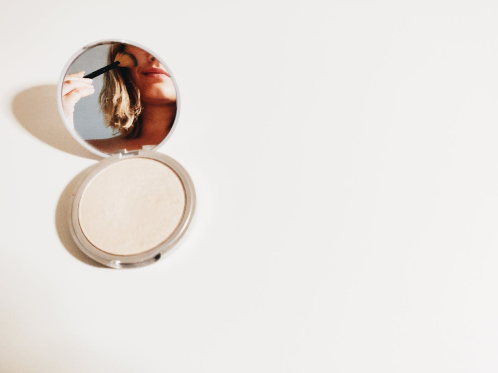 Close-up of a woman using makeup in a compact mirror, set against a bright background. k-beauty sverige/danmark/norge
