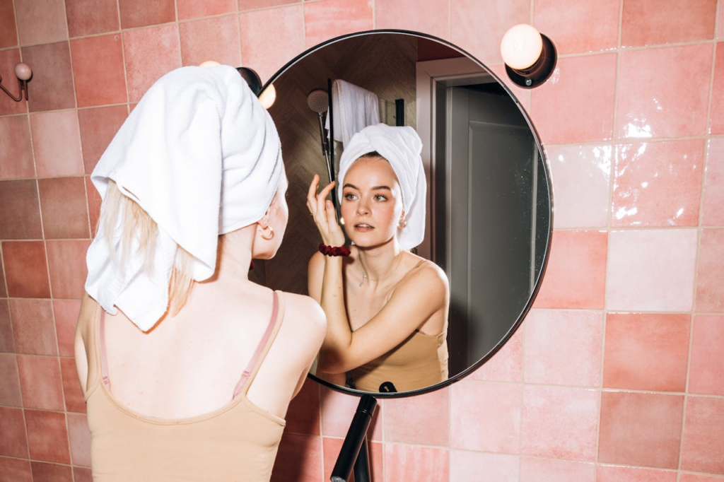 A young woman with a towel in a pink bathroom, reflecting in round mirror as she performs skincare.