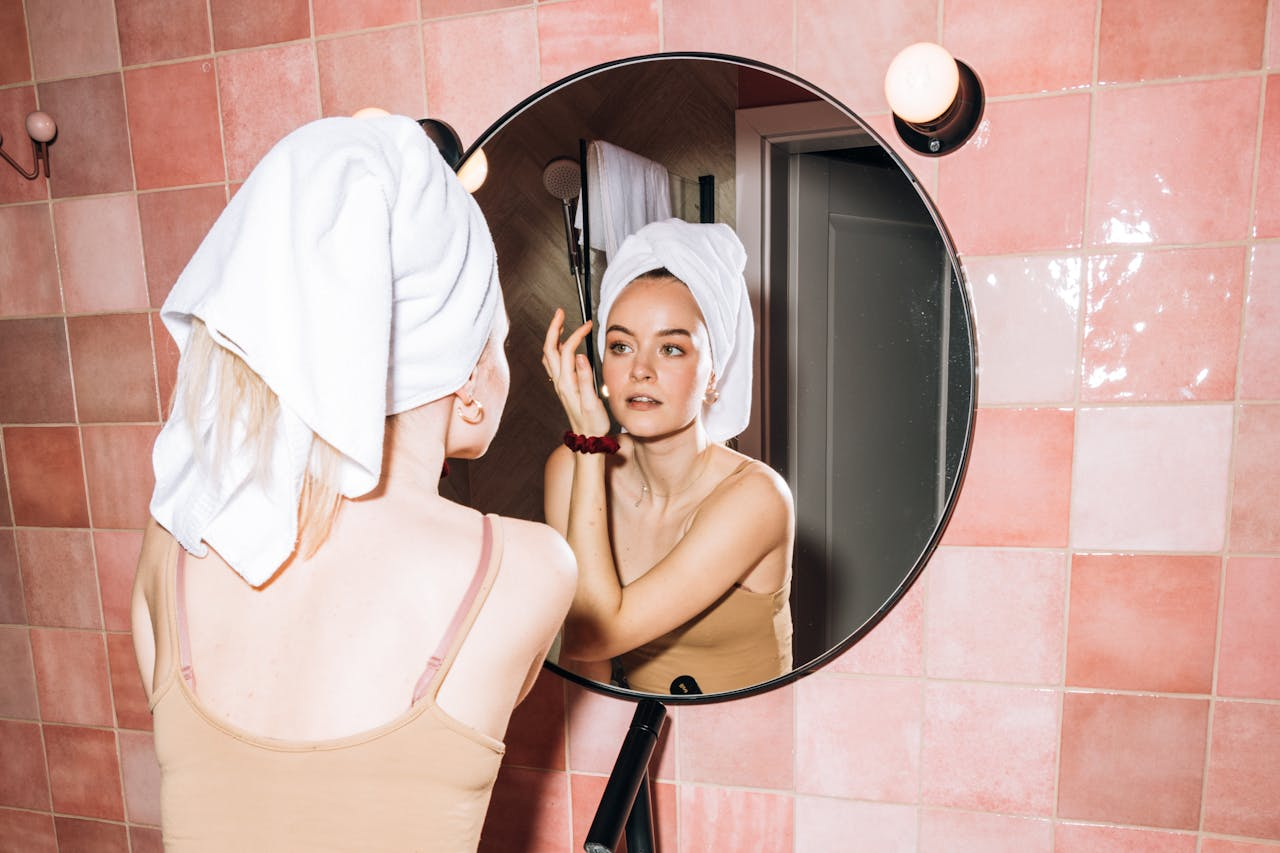 A young woman with a towel in a pink bathroom, reflecting in round mirror as she performs skincare.